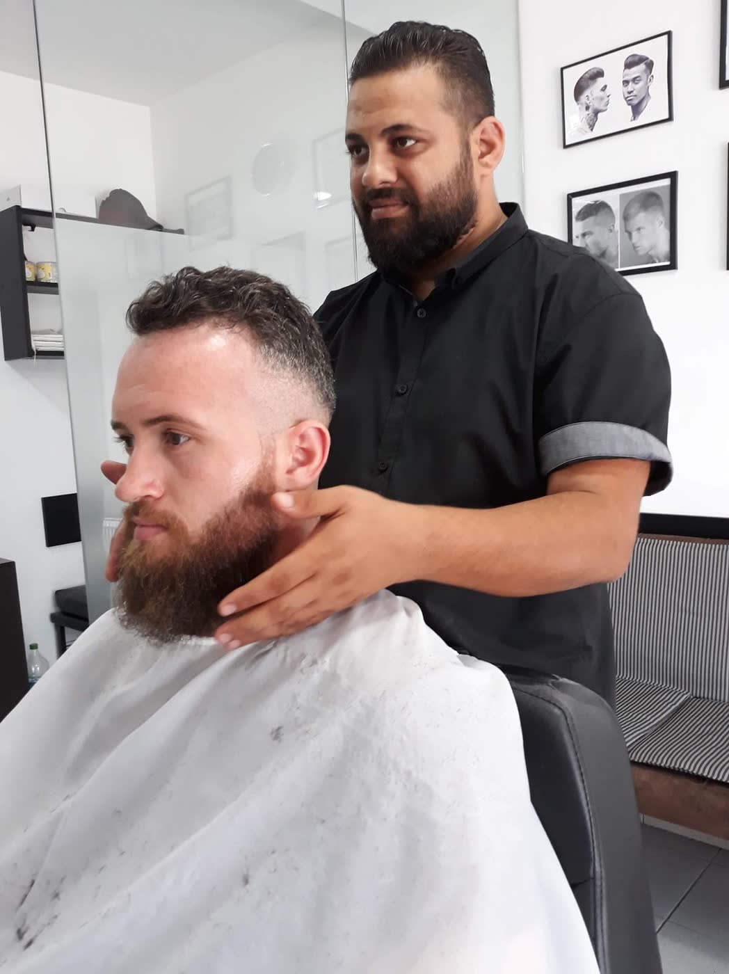 Frizerul, în cămașă neagră, stă în picioare în spatele scaunului și îi prinde cu ambele mâini barba roșcată unui client cu pelerină albă