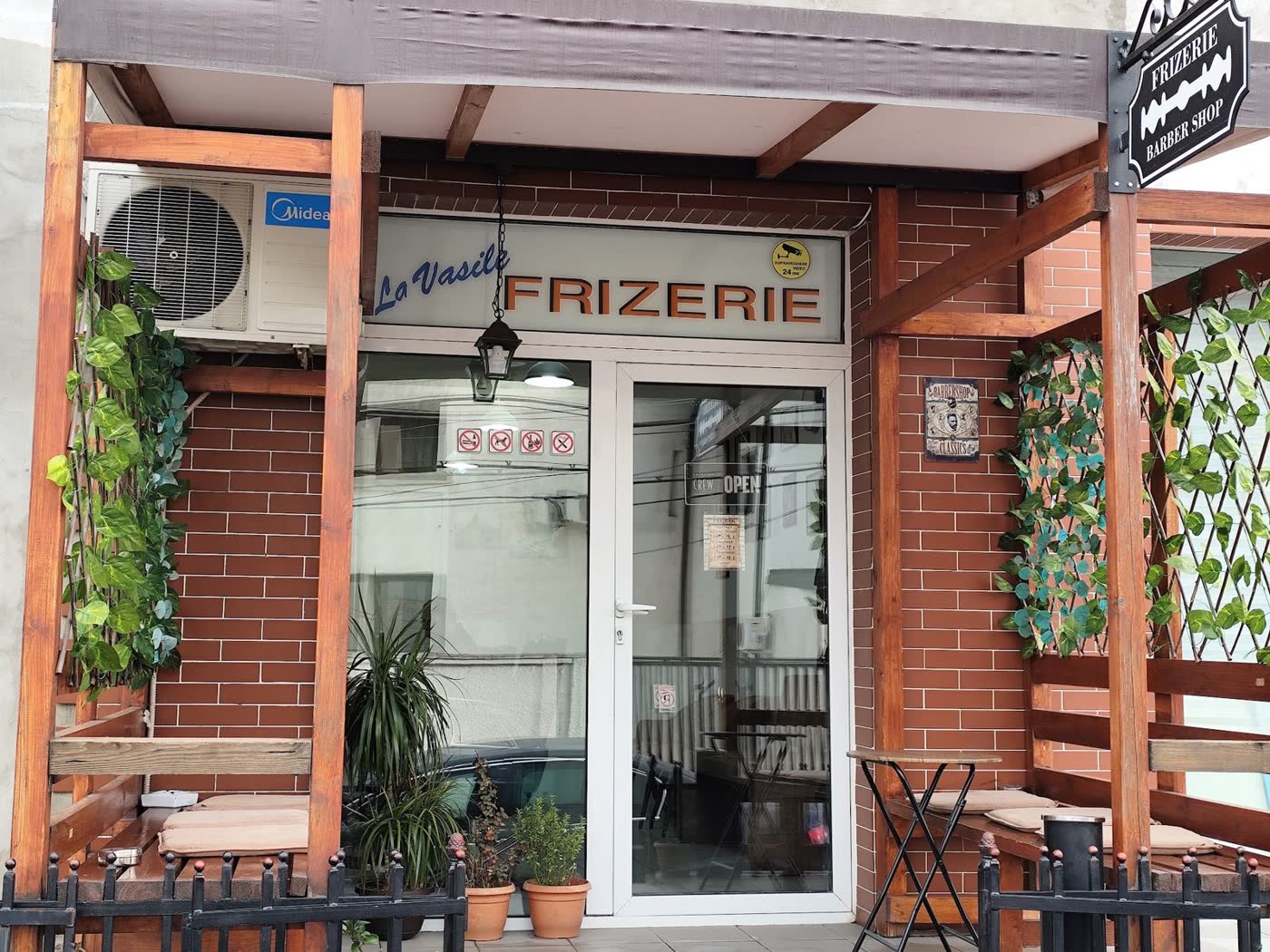 Fațada frizeriei: cărămidă, pergolă de lemn, firma La Vasile Frizerie și plante verzi