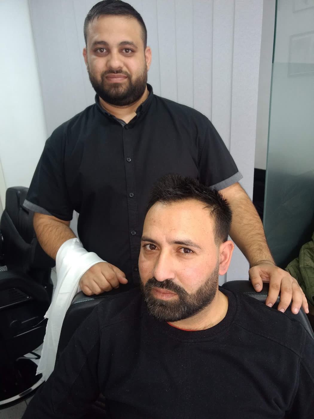 Frizerul în cămașă neagră, în picioare în spatele unui client așezat, cu barba proaspăt conturată